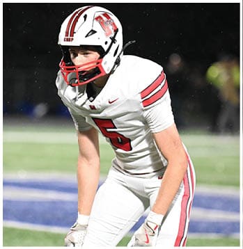 A person wearing a white football uniform with red stripes and the number five is bent forward on a football field, appearing ready to play. Description generated by AI
