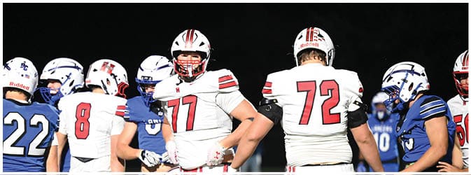 Adults wearing football uniforms from two different teams are standing on a field at night, appearing to prepare for or discuss the game. Description generated by AI