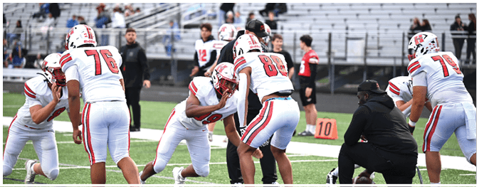 People in football uniforms are practicing on a field with a coach kneeling nearby and others standing in the background. Description generated by AI