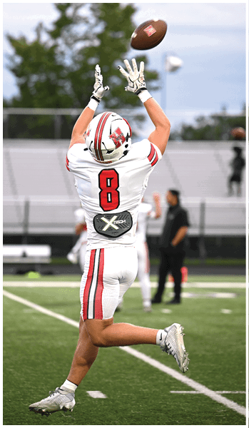 A person in a white football uniform with the number eight is jumping with arms raised to catch a football on a field with blurred spectators in the background. Description generated by AI