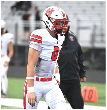 A person wearing a white and red football uniform stands on a field near an orange yard marker with the number 10. Description generated by AI