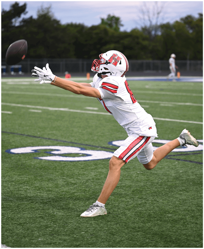 A person in a white football uniform with red and black stripes is reaching out with both hands to catch a football on a green field. Description generated by AI