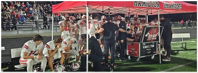 People in football uniforms and coaches are gathered under a red tent labeled Hamilton Chargers on a sports field with spectators in the background. Description generated by AI