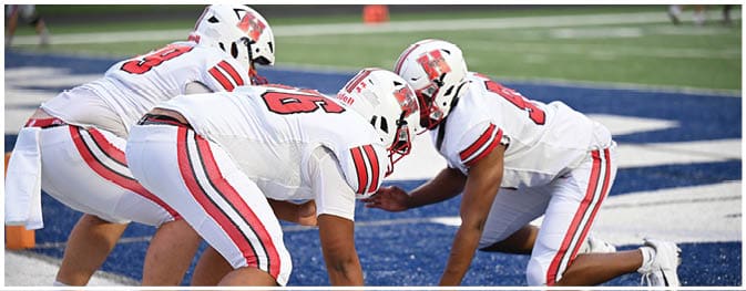 Two people in white football uniforms with red and black stripes are crouched on a blue field, facing each other in a ready position. Description generated by AI