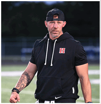 A person wearing a black sleeveless hoodie, black cap worn backward, and black pants is standing on a sports field holding a clipboard and a football. Description generated by AI