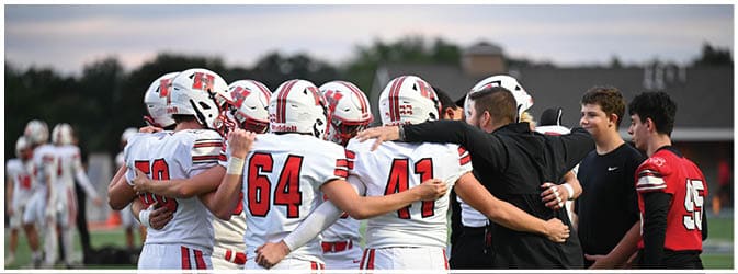 A group of people in football uniforms and coaches are huddled together on a field while two people in different colored jerseys stand nearby. Description generated by AI
