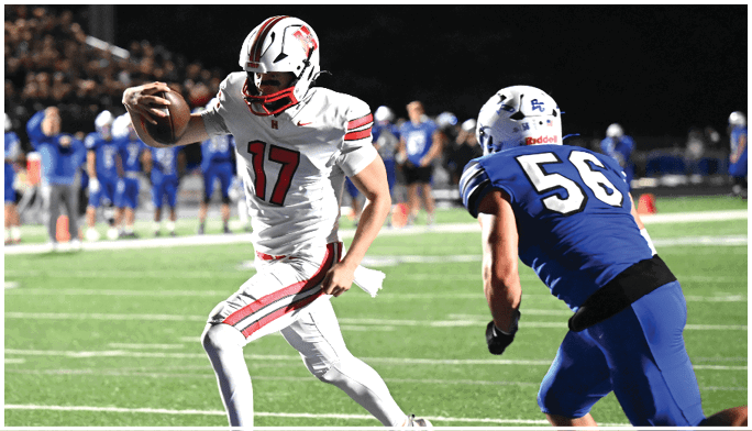 A person in a white football uniform is running with the ball while another person in a blue football uniform is chasing them on a lit field at night. Description generated by AI