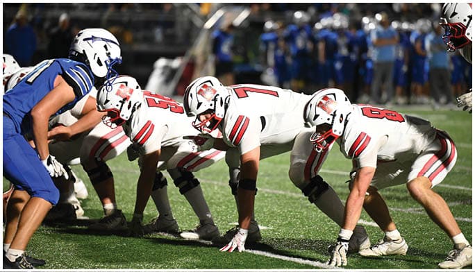 Players in football uniforms from two opposing teams are lined up facing each other on a field, preparing for a play during a game. Description generated by AI