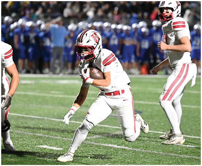 Two people in white football uniforms with red stripes are playing on a green field with a crowd and players in blue uniforms in the background. Description generated by AI