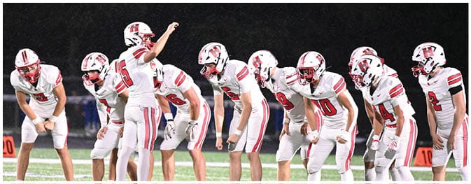 A group of people in white football uniforms and helmets are lined up on a field at night, with one person raising an arm as if giving a signal. Description generated by AI