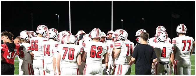 A group of people wearing white football uniforms with red stripes and numbers are gathered on a field at night. Description generated by AI