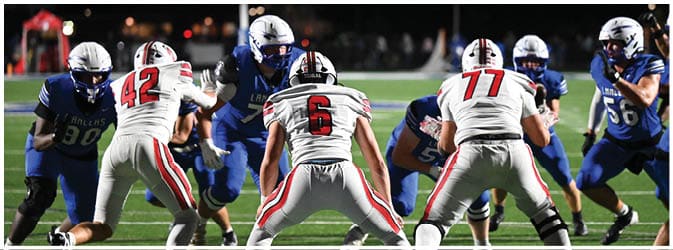 People in football uniforms are lined up on a field preparing for a play under stadium lights at night. Description generated by AI