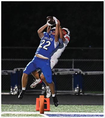 Two people in blue football uniforms and one person in a white uniform are jumping to catch a football near the end zone on a lit field at night. Description generated by AI
