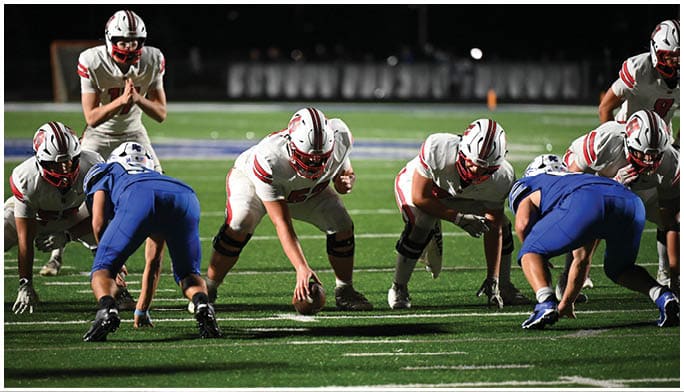 People in athletic clothing are positioned on a football field preparing for a play, with one person holding a football. Description generated by AI