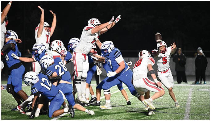 Players in blue and white uniforms are engaged in a football play on a field under bright lights with one player in white throwing the ball. Description generated by AI