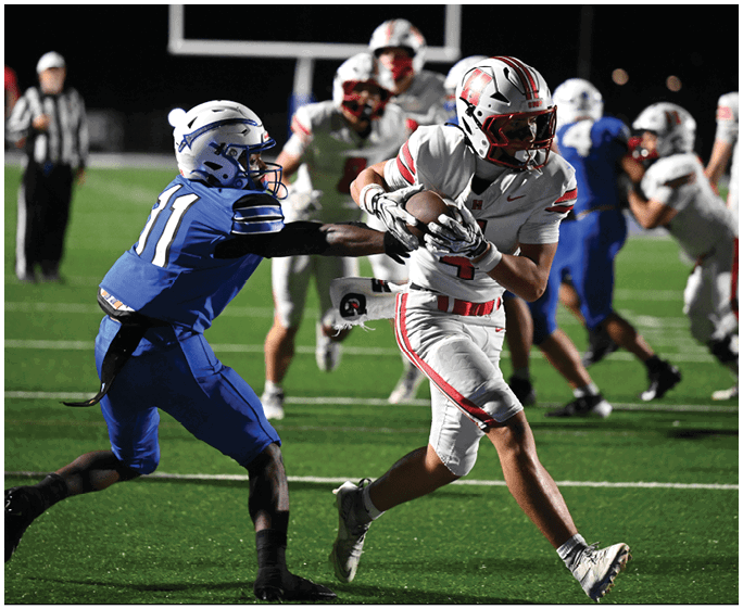 A person in a white and red football uniform is running with the ball while another person in a blue uniform attempts to grab their arm on a football field. Description generated by AI