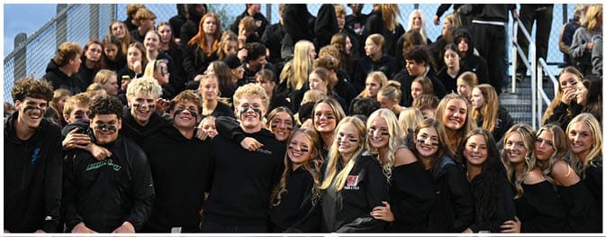 A large group of people wearing black clothing are gathered closely together on bleachers, smiling and posing for a photo in an outdoor setting. Description generated by AI