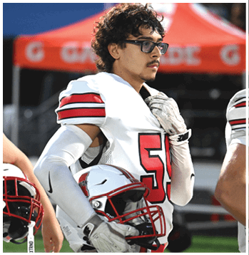 A person wearing a white football uniform with the number 55 and glasses stands on a field holding a helmet with a red and black stripe, with a red tent and blurred crowd in the background. Description generated by AI