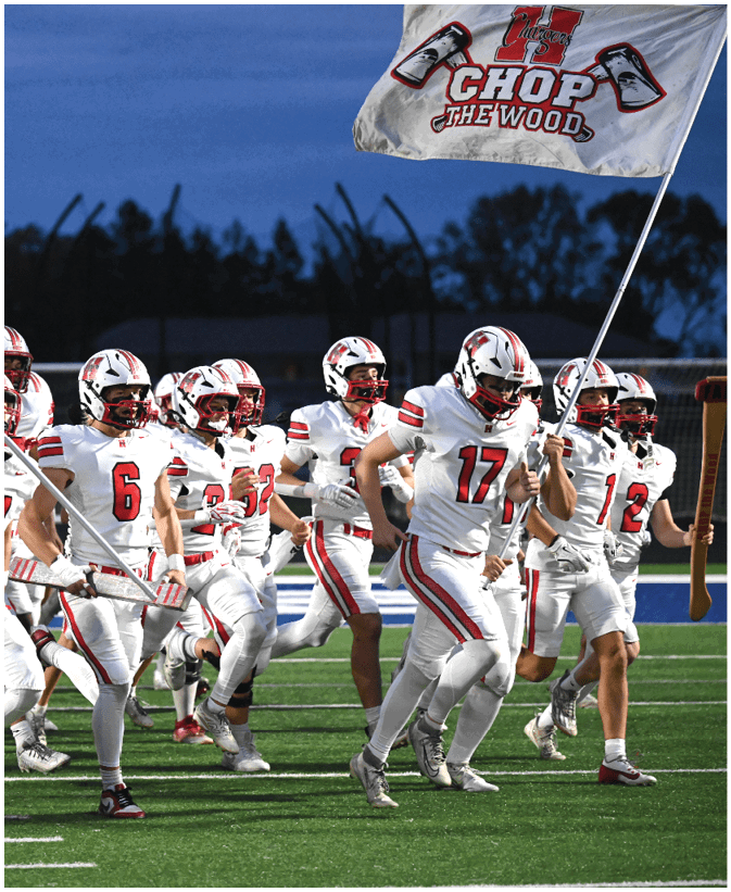 A group of football players in white uniforms with red stripes are running on a field at dusk, with one player holding a flag. Description generated by AI