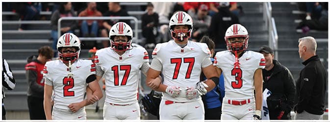 Four football players in white uniforms stand arm in arm on a field with a referee and coaches nearby and spectators in the background. Description generated by AI