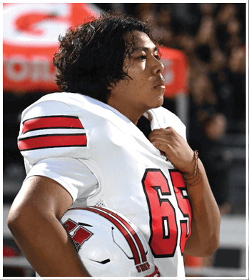 A person wearing a white football jersey with the number 65 is holding a white helmet with red and black stripes while looking to the side. Description generated by AI