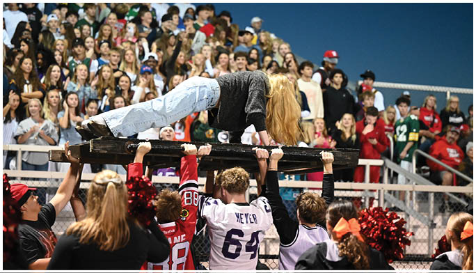 A group of people in sports jerseys are holding up a person lying horizontally on a wooden plank above their heads in a crowded outdoor event. Description generated by AI