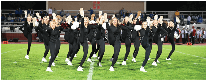 A group of adults dressed in black athletic outfits perform a high kick routine on a football field at night with pom-poms in hand. Description generated by AI