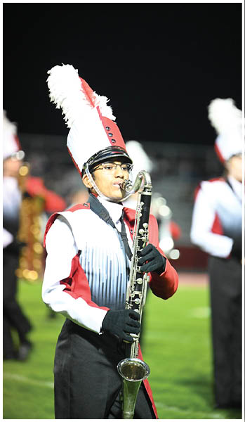 A person in a red, white, and black marching band uniform with a tall feathered hat is playing a clarinet on a grassy field at night. Description generated by AI