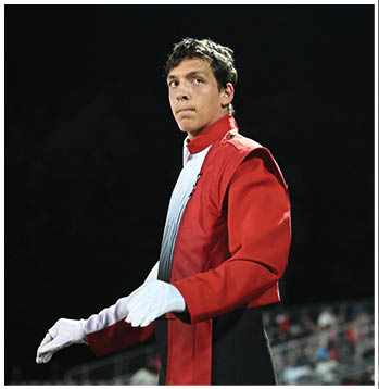 A person wearing a red and white marching band uniform looks thoughtfully to the side against a dark background. Description generated by AI