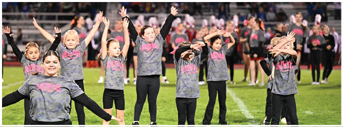 Children wearing matching shirts are performing a coordinated dance or cheer routine on a field with people and bleachers in the background. Description generated by AI