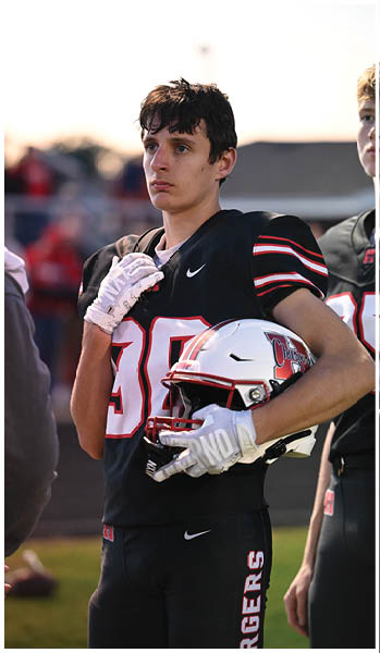 A person in a black football uniform with the number 30 holds a white helmet and appears focused while standing on a field during a game. Description generated by AI