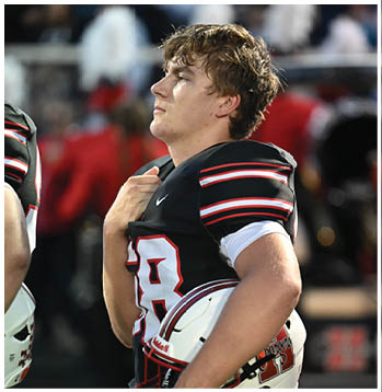 A person wearing a black football jersey with red and white stripes is standing on a field, holding their helmet and looking to the side. Description generated by AI
