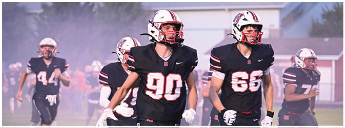 Five people wearing black football uniforms with helmets are running on a field with a misty background. Description generated by AI