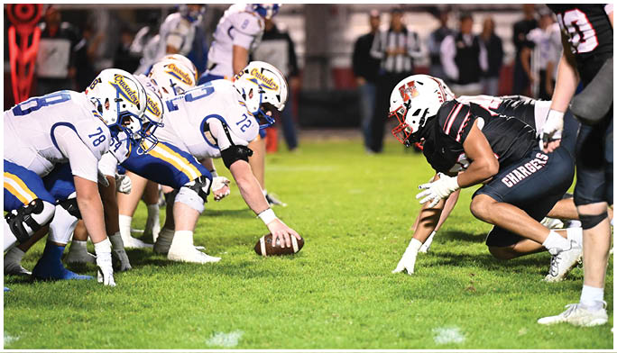 Two opposing American football teams are lined up at the line of scrimmage on a grassy field during a nighttime game. Description generated by AI