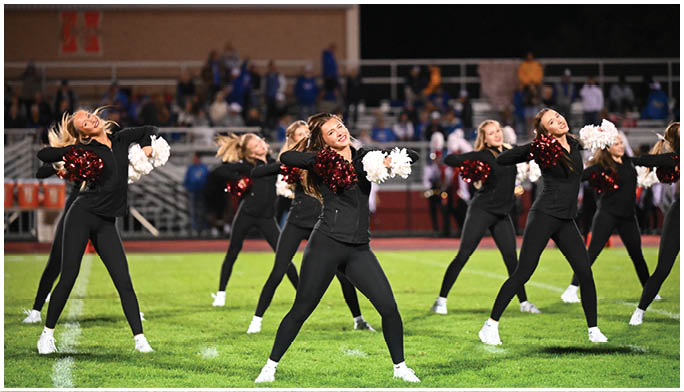 A group of adults dressed in black athletic outfits perform a synchronized cheerleading routine on a grassy field at night with a crowd in the background. Description generated by AI