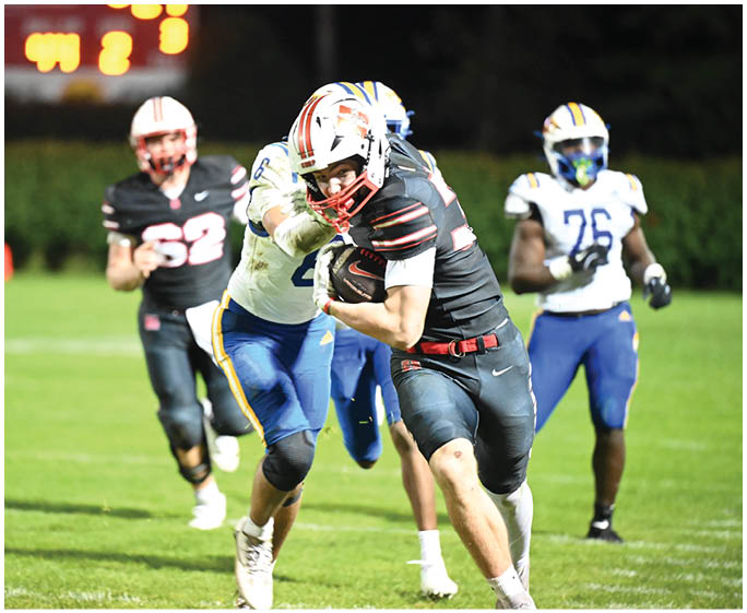 Two people in football uniforms are engaged in a play on a grassy field with one person holding the ball and another attempting to tackle. Description generated by AI