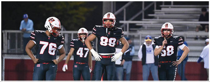 Five people wearing football uniforms and helmets stand on a field with a blurred background of spectators and staff. Description generated by AI