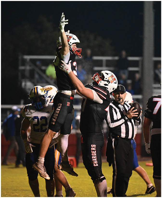 Players in football uniforms are engaged in a game on a field under stadium lights with one player jumping to catch the ball while others watch or prepare to react. Description generated by AI