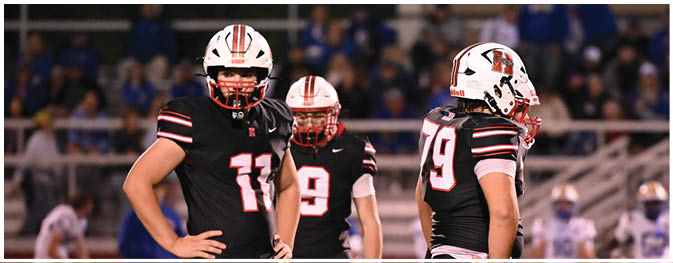 Three football players in black uniforms and helmets stand on the field near a referee holding a football and pointing during a game. Description generated by AI