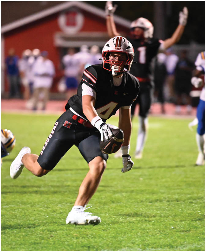 A person wearing a black football uniform with the number four is running on a grassy field while holding a football. Description generated by AI