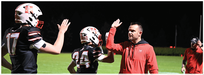 A person in a red jacket is giving a high five to a football player wearing a helmet and jersey number 24 on a field at night. Description generated by AI