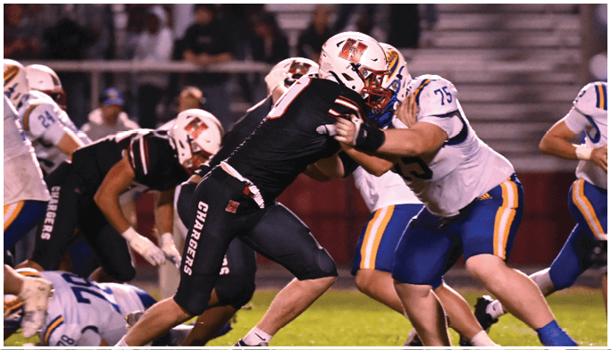 Players in football uniforms are engaged in a game on a grassy field with some players blocking and others tackling. Description generated by AI