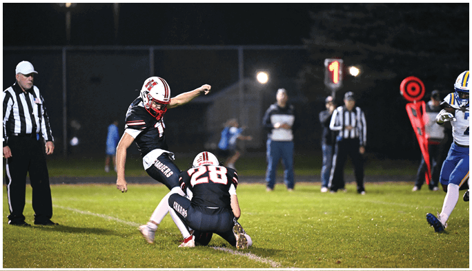 A person in a black and red football uniform is kicking a football held by another person kneeling on the grass field during a nighttime game with referees and other players in the background. Description generated by AI
