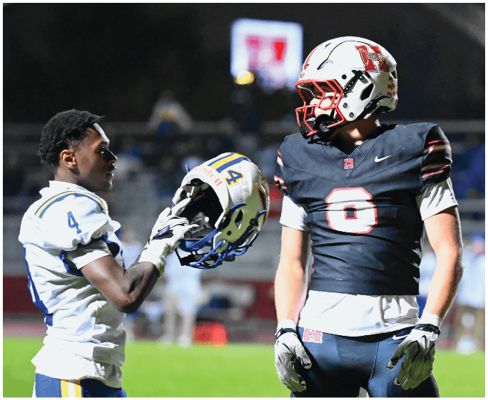 Two people in football uniforms face each other on a field, with one holding a helmet and the other wearing a helmet. Description generated by AI