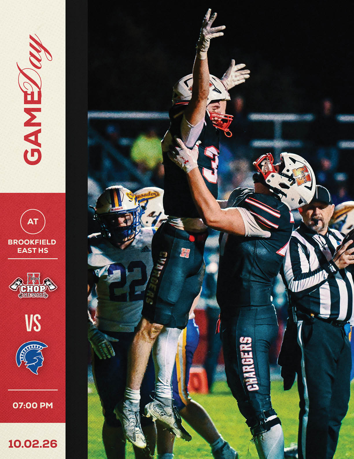 Two football players in dark uniforms celebrate on the field with one player lifting the other while a referee and opposing players in white and blue uniforms stand nearby during a night game. Description generated by AI
