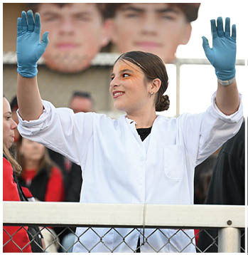A person wearing a white lab coat is standing behind a chain-link fence with arms raised, with blurred people in the background. Description generated by AI