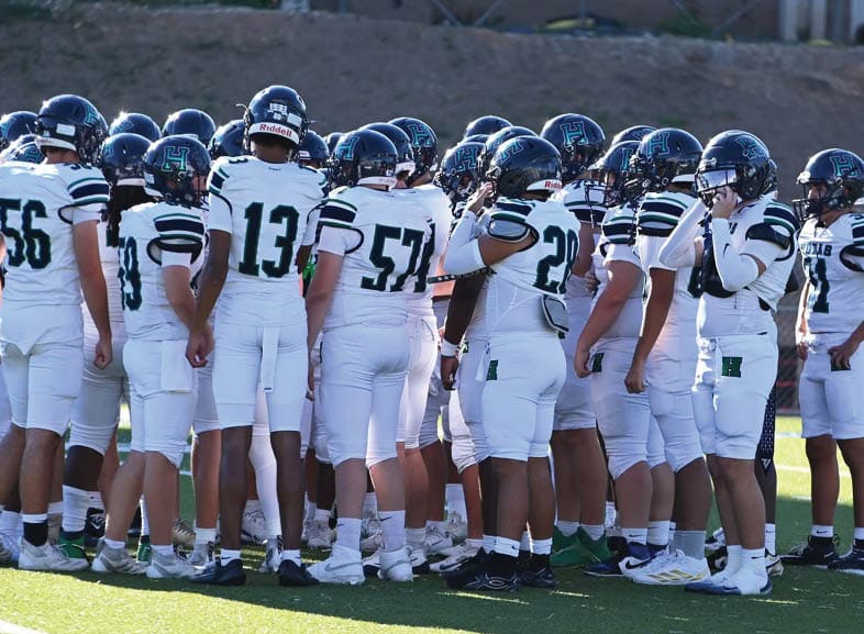 Four people wearing black football uniforms with “VISTA" on the front are standing together in a stadium with a crowd in the background, two of them holding helmets raised above their heads. Description generated by AI