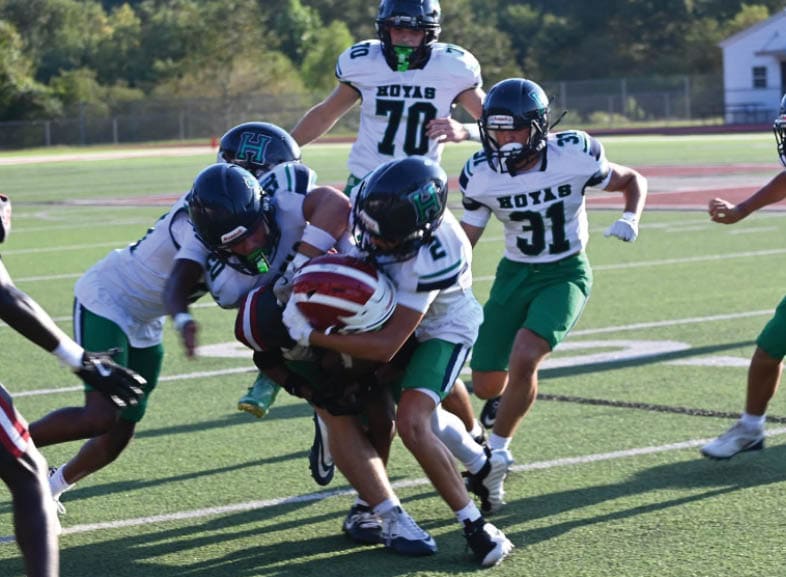 Four people wearing black football uniforms with “VISTA" on the front are standing together in a stadium with a crowd in the background, two of them holding helmets raised above their heads. Description generated by AI