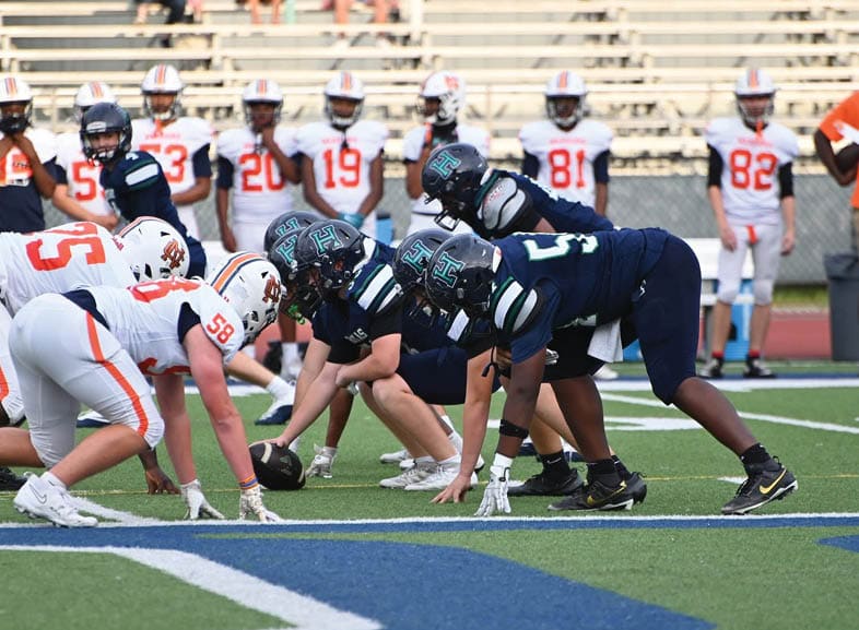 Four people wearing black football uniforms with “VISTA" on the front are standing together in a stadium with a crowd in the background, two of them holding helmets raised above their heads. Description generated by AI