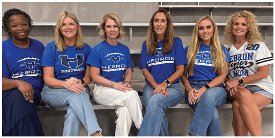 Six adults sitting on bleachers wearing casual clothes and matching blue shirts with “Hebron" and football-themed designs, smiling and posing for the photo. Description generated by AI